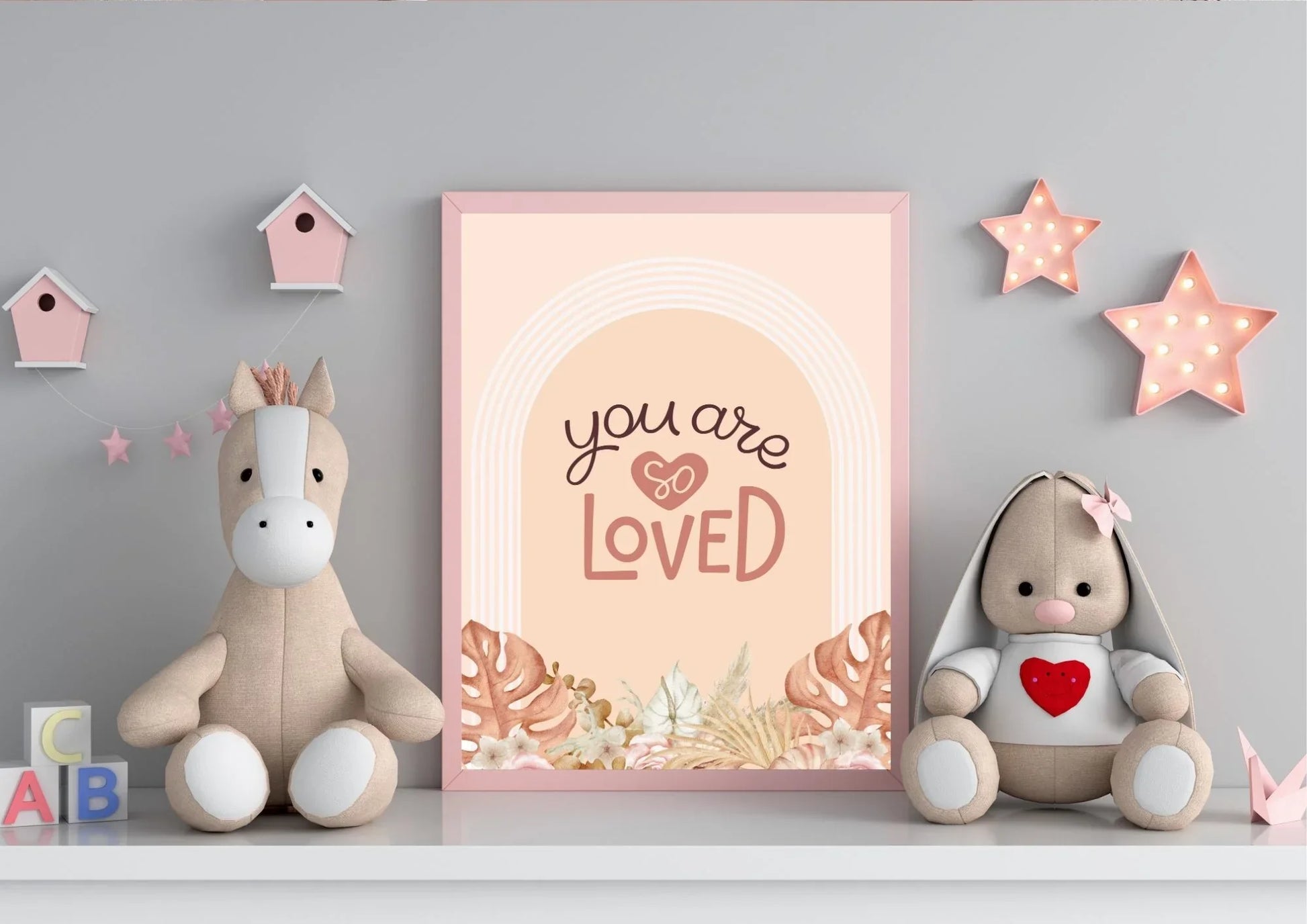 
A framed "You are so LOVED" boho art print on a shelf with stuffed animals and pink star lights.
