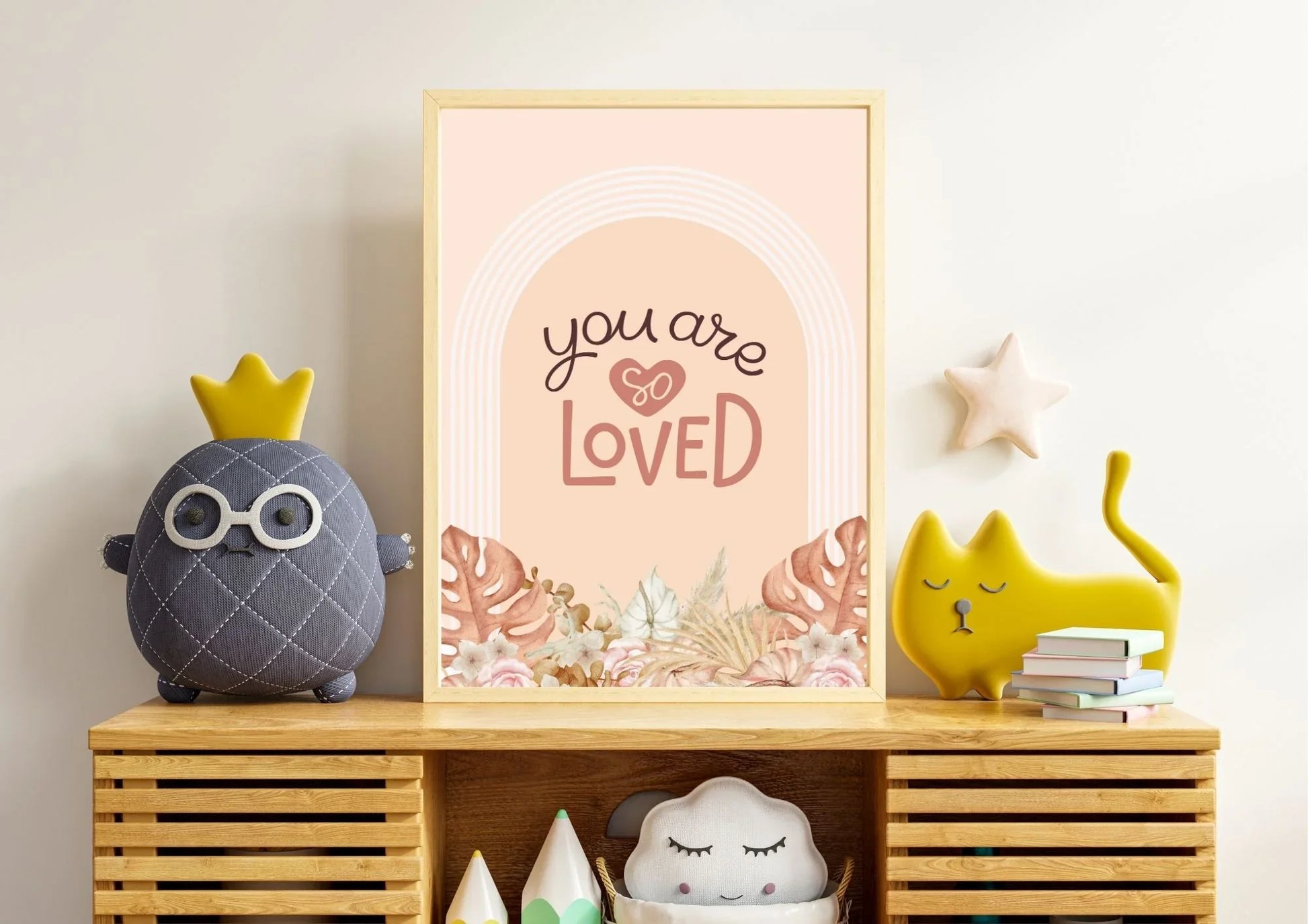 A wooden-framed "You are so LOVED" boho art print on a cabinet with a variety of whimsical pillows.
