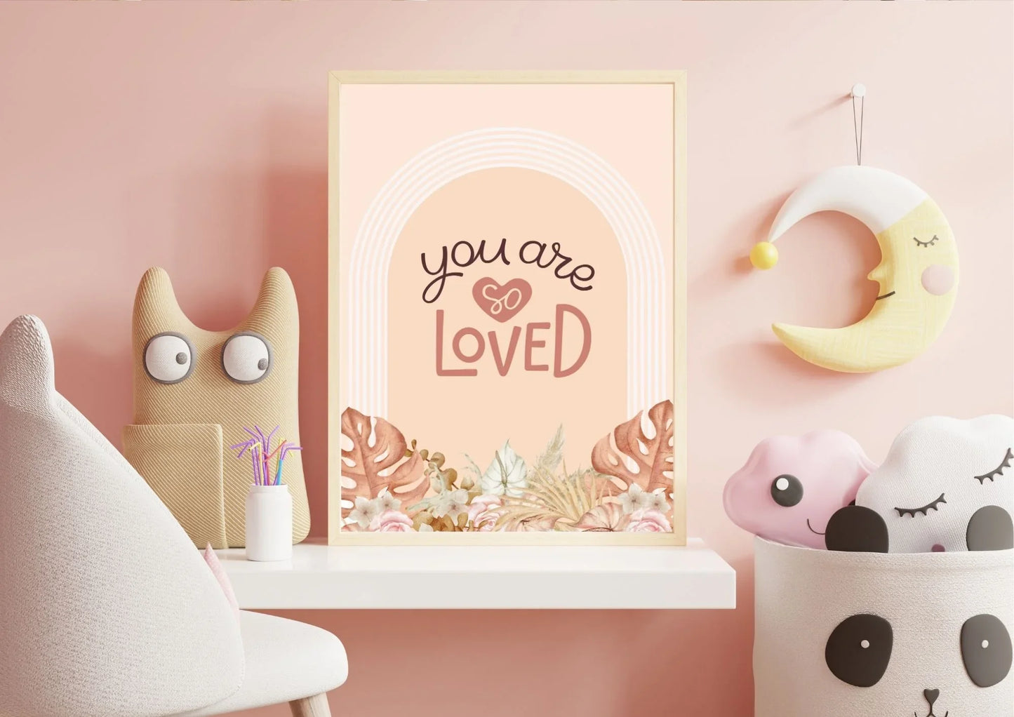 A wooden-framed "You are so LOVED" boho art print on a shelf in a child's bedroom with a sleepy moon and stuffed toys.