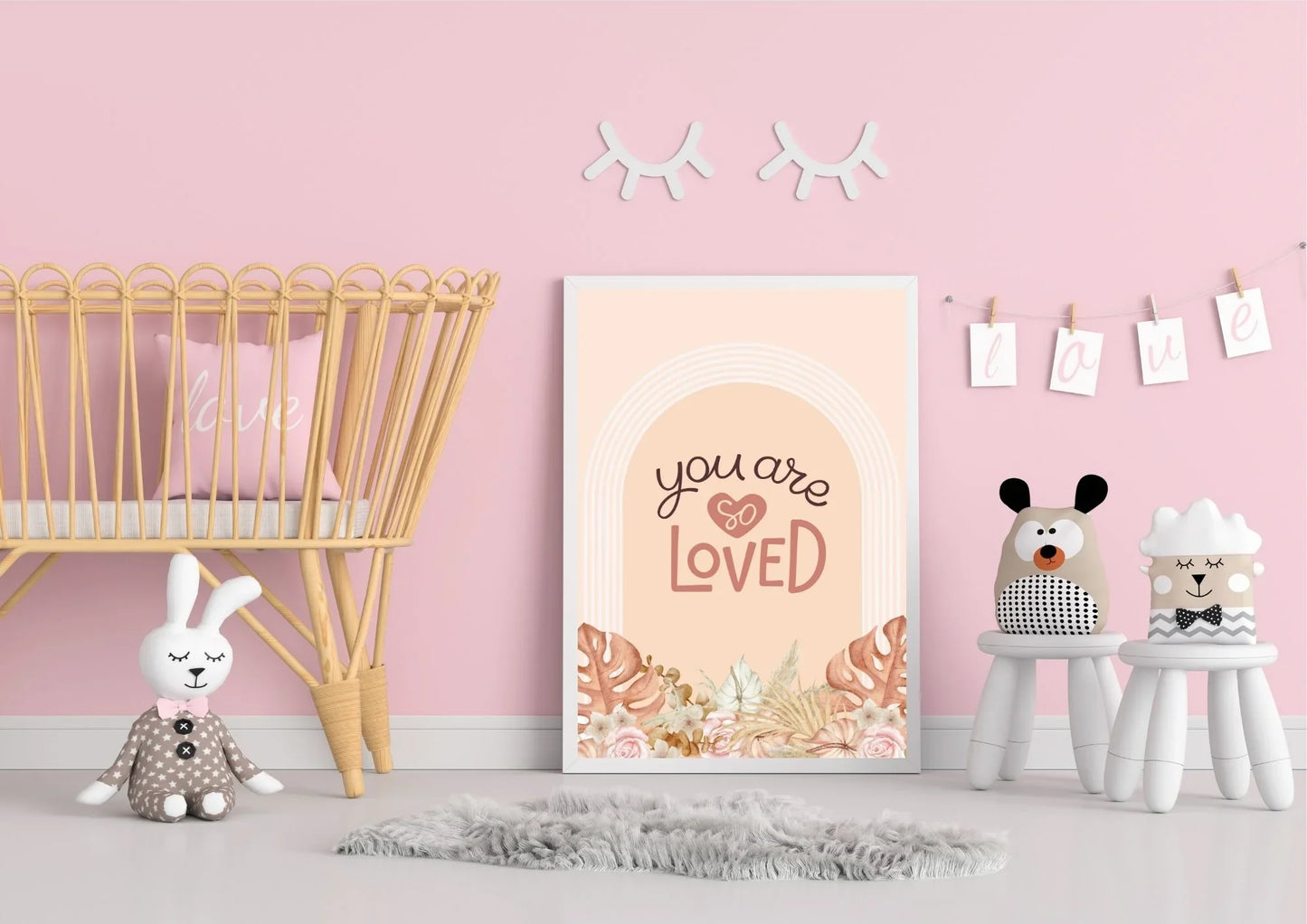 A framed "You are so LOVED" boho art print on the floor of a nursery with a wicker crib and a gray rug.