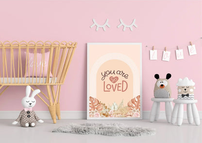 A framed "You are so LOVED" boho art print on the floor of a nursery with a wicker crib and a gray rug.