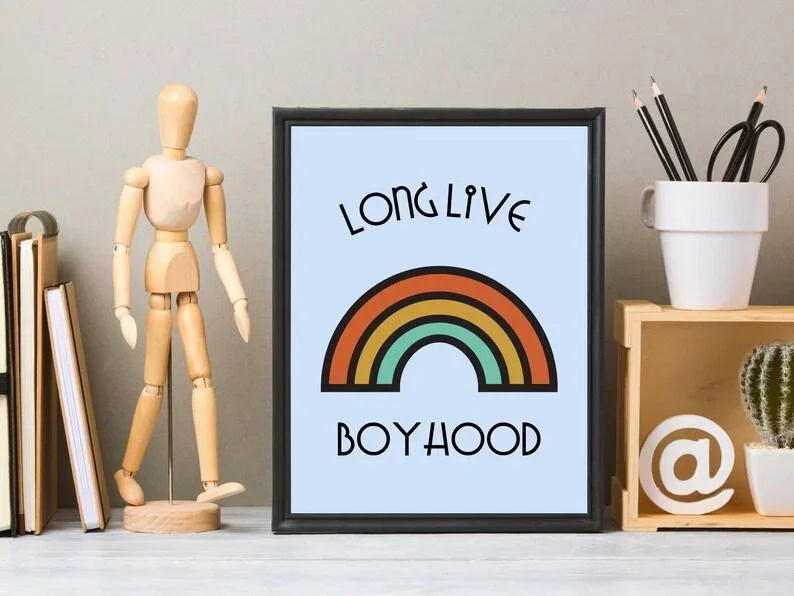 A close-up of a framed "Long Live Boyhood" retro rainbow print on a desk, next to a wooden mannequin, books, and a pencil holder.