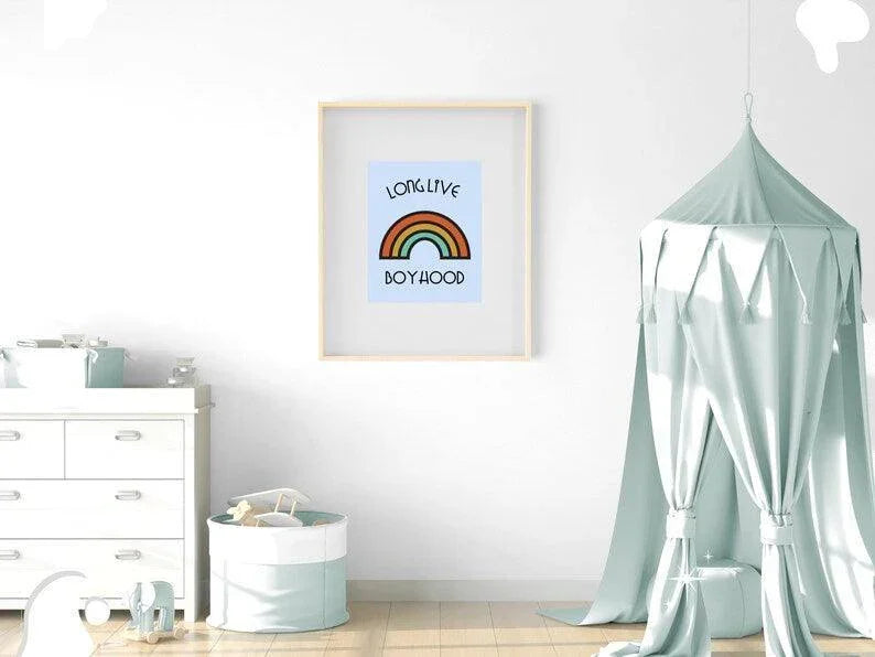 
A framed "Long Live Boyhood" retro rainbow print hanging on a white wall in a neutral-toned nursery, above a changing table and next to a bed with a canopy.