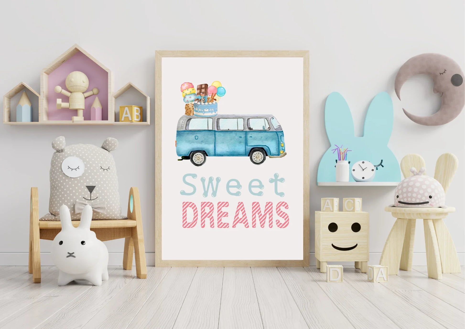 A framed art print is centered on the white floor against a white wall in a child's playroom. The artwork is a watercolor illustration of a blue, vintage Volkswagen van (VW bus) with a stack of ice cream cones, popsicles, and a birthday cake on its roof, and the text "Sweet DREAMS" in light blue and striped pink below it