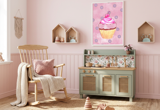 A photorealistic photograph of a cozy boho-themed nursery during a bright morning. The room features a soft, light pink wall with wainscoting and a light oak wood floor partially covered by a large round jute rug. In the center, a sage green play kitchen with wicker-inlay doors, light wood countertops, and a floral backsplash sits against the wall. To the left is a natural wood rocking chair with a cream-colored knit throw blanket and a dusty pink velvet pillow. Hanging prominently above the kitchen is a wh