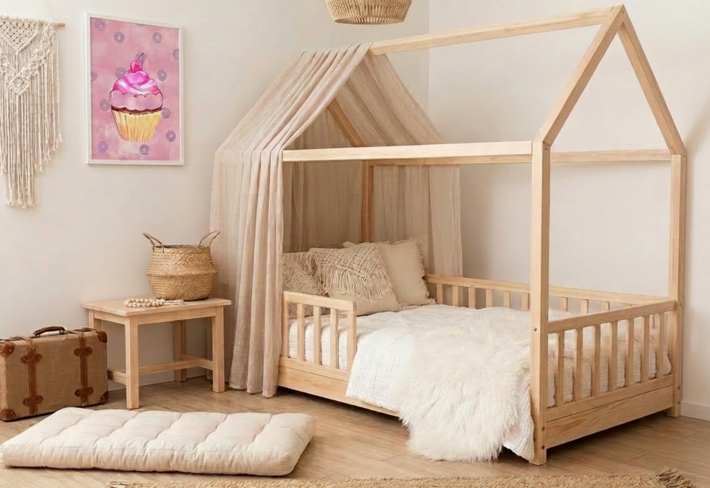 A photorealistic photograph of a neutral, boho-themed toddler bedroom featuring a light natural wood house-shaped floor bed. The bed includes a low slatted railing and is styled with a white fluffy throw blanket and cream-colored textured pillows under a beige sheer canopy. Hanging on the cream wall to the left is a white-framed vertical art print of a whimsical pink watercolor cupcake with purple donuts on a pink background. A small wooden side table sits next to the bed holding a woven basket and a wooden