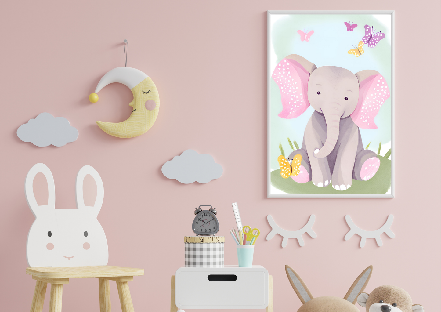 A framed digital art print of a light gray baby elephant with pink ears and feet, sitting in a field of grass with butterflies. The print hangs on a pink wall in a nursery, with a crescent moon and cloud decor to the left.