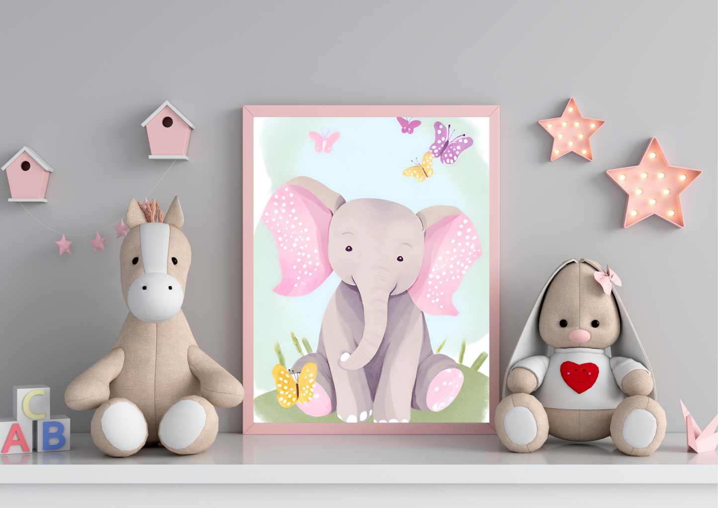 A framed digital art print of a baby elephant with pink ears and butterflies, leaning against a gray wall on a white shelf. A light brown horse plush toy is on the left, and a beige stuffed rabbit with a red heart on its chest is on the right. Pink star-shaped lights are hanging on the wall.