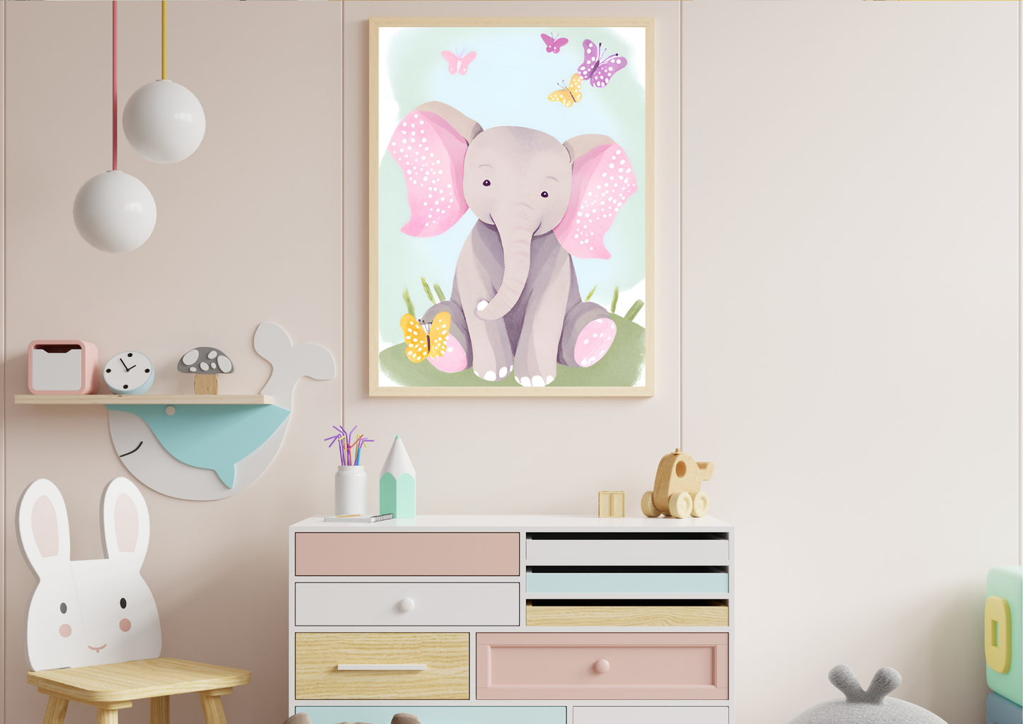 A framed digital art print of a baby elephant with pink ears and butterflies, hanging on a light pink wall. The print is positioned above a colorful dresser with multiple drawers and a small whale-shaped shelf with toys.