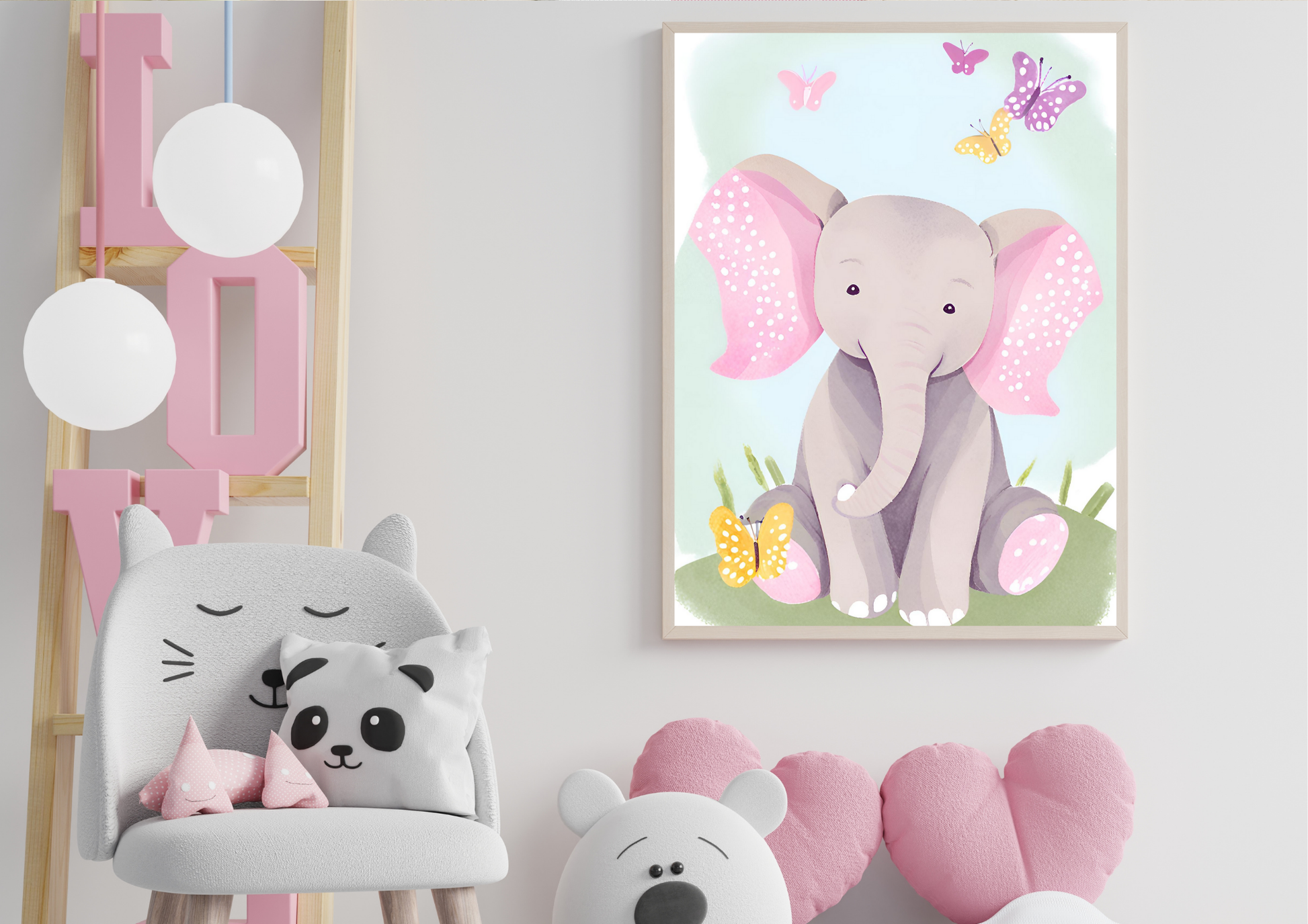 A framed digital art print of a baby elephant with pink ears and butterflies, hanging on a white wall. To the left, a light wood ladder with a large pink letter "L" is visible, along with a white cat-shaped chair and pillows. In the foreground, a stuffed teddy bear and two pink heart pillows are on the floor.