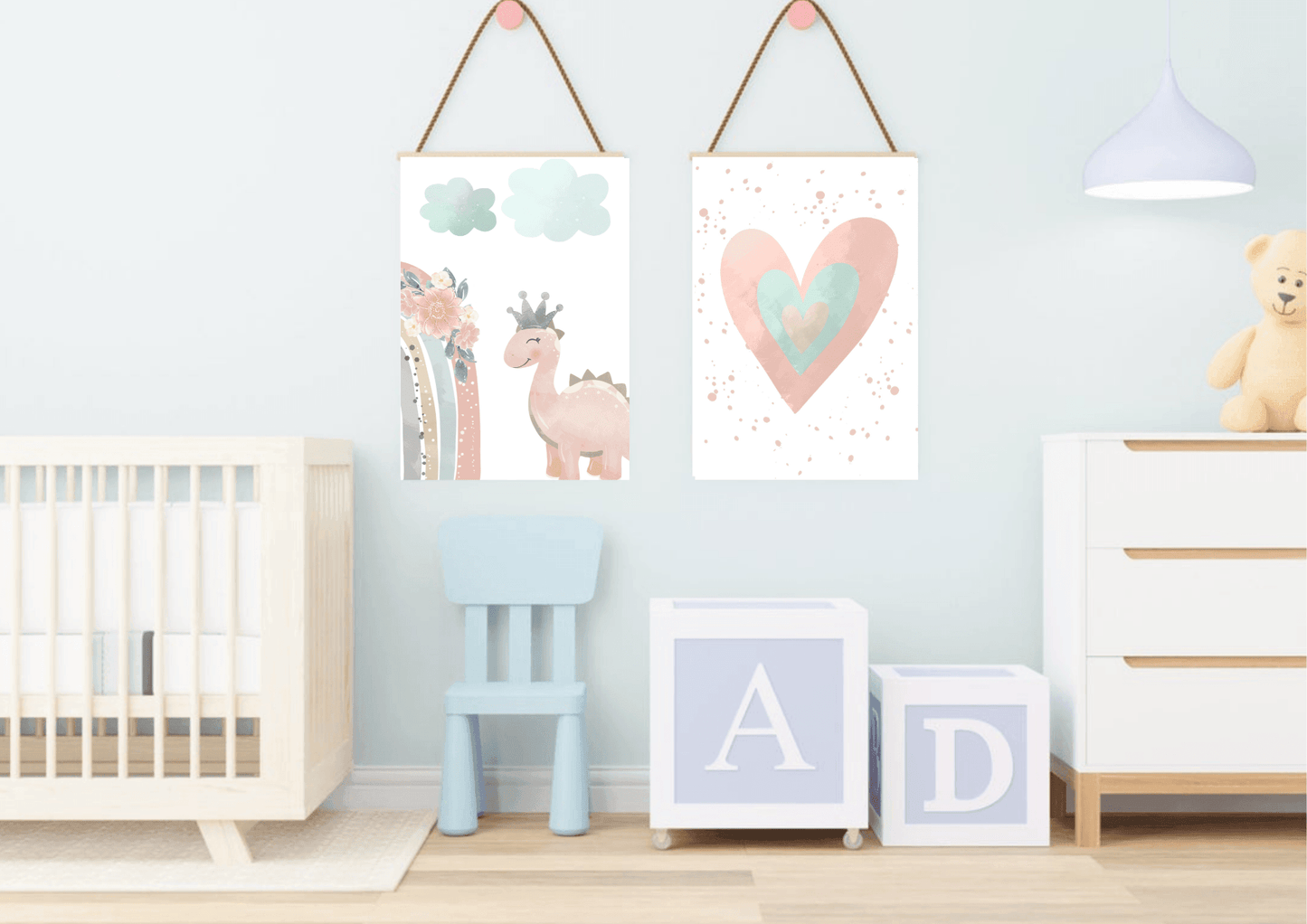 A playful baby's room with a light blue wall. Two framed prints hang from the wall with rope, showing a cute pink dinosaur with a crown and a watercolor heart. The room also features a crib, a small blue chair, and large letter blocks, creating a sweet and inviting space.