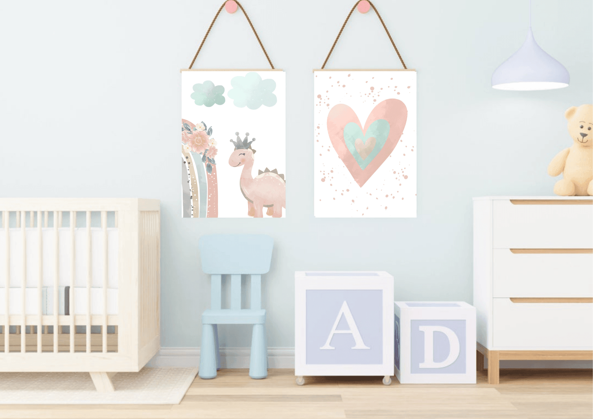 A playful baby's room with a light blue wall. Two framed prints hang from the wall with rope, showing a cute pink dinosaur with a crown and a watercolor heart. The room also features a crib, a small blue chair, and large letter blocks, creating a sweet and inviting space.