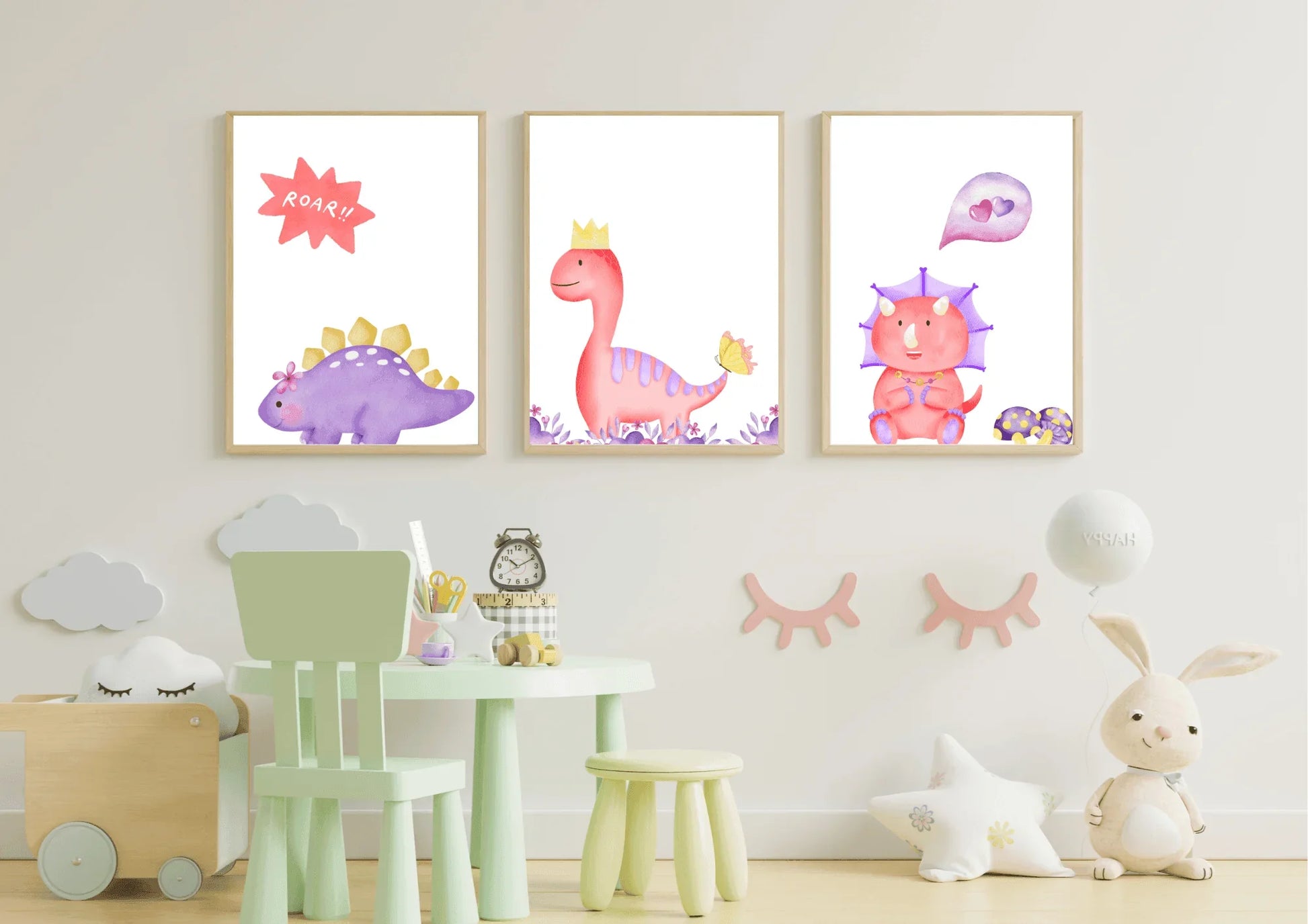 A trio of framed digital art prints hanging on a white wall in a child's room. The prints feature three watercolor dinosaurs: a purple stegosaurus with a speech bubble that says "ROAR!!", a pink T-Rex with a crown, and a pink triceratops with a heart bubble. Below the prints is a small mint green table and chair set.
