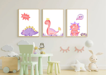 A trio of framed digital art prints hanging on a white wall in a child's room. The prints feature three watercolor dinosaurs: a purple stegosaurus with a speech bubble that says "ROAR!!", a pink T-Rex with a crown, and a pink triceratops with a heart bubble. Below the prints is a small mint green table and chair set.