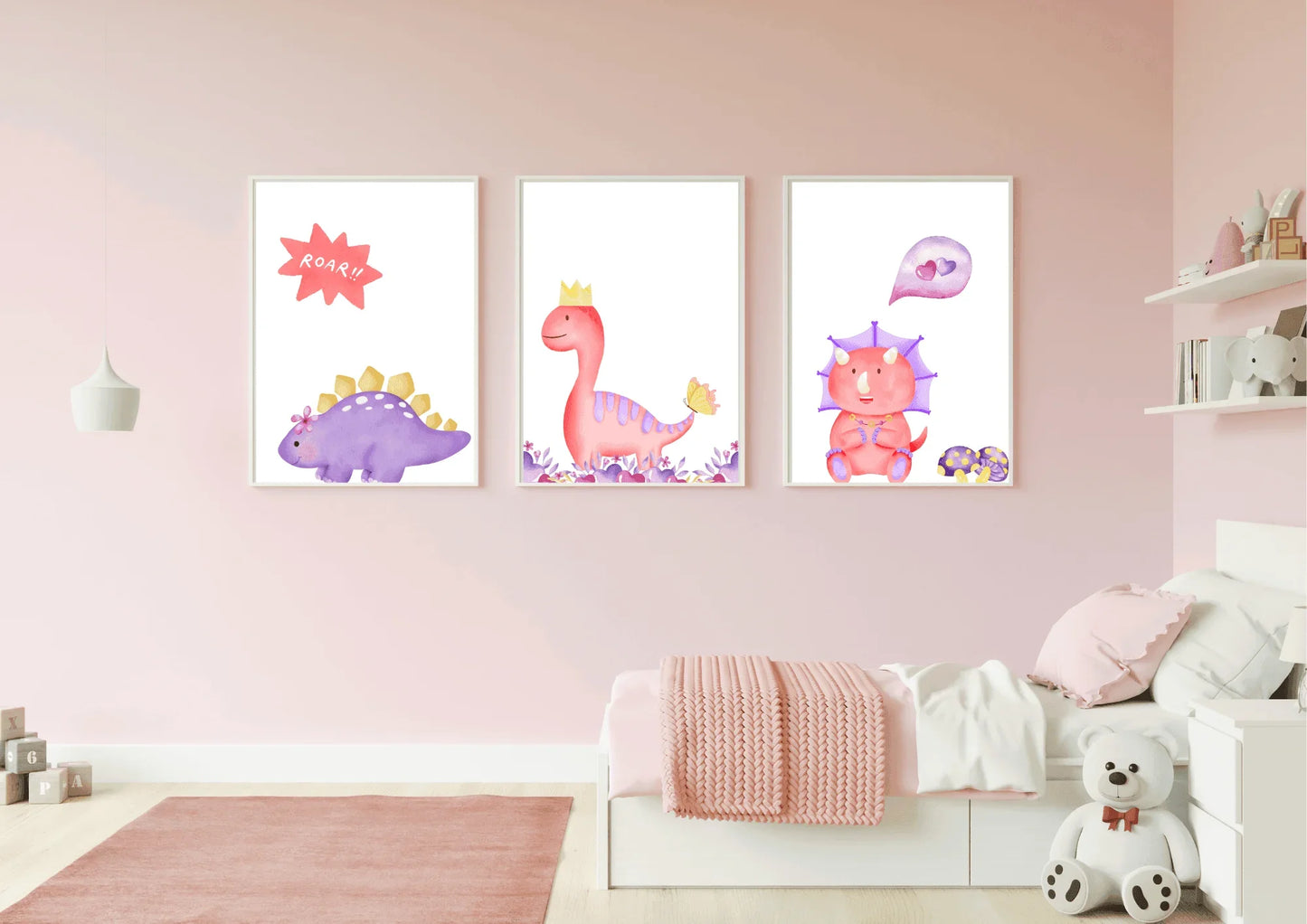 A set of three framed digital art prints featuring watercolor dinosaurs, hanging on a light pink wall above a small bed with a pink rug. The prints show a purple stegosaurus, a pink T-Rex with a crown, and a pink triceratops with a heart bubble. A white teddy bear sits on the floor next to the bed.