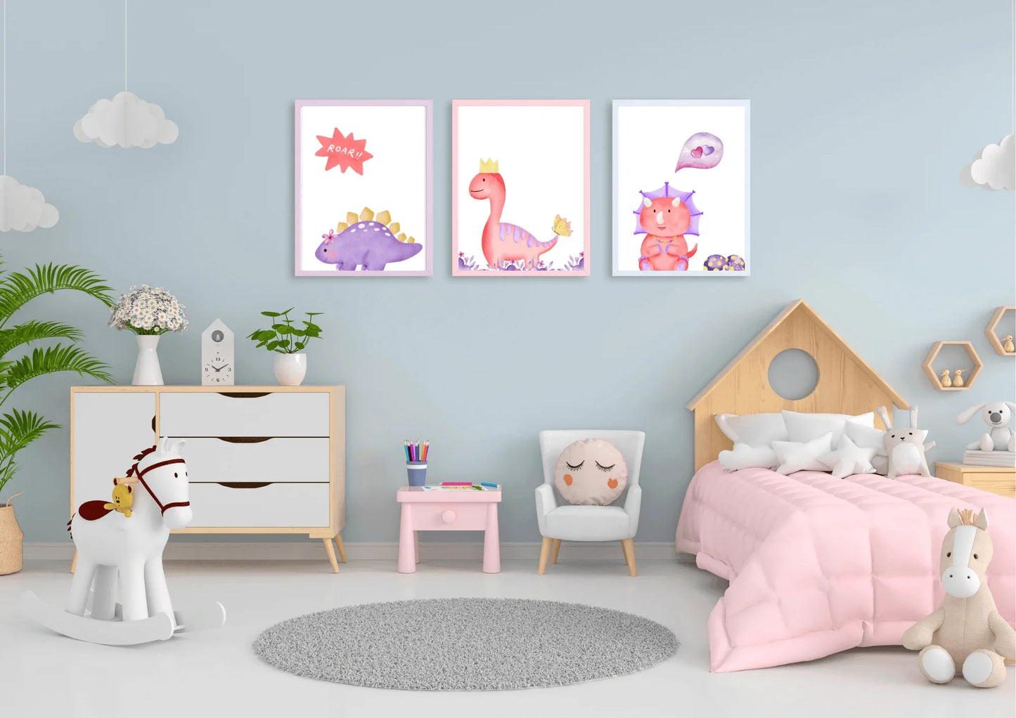 
A trio of framed digital art prints featuring watercolor dinosaurs, hanging on a light blue wall in a child's bedroom. The prints show a purple stegosaurus, a pink T-Rex with a crown, and a pink triceratops with a heart bubble. The room is decorated with a small dresser, a rocking horse, and a bed shaped like a house.