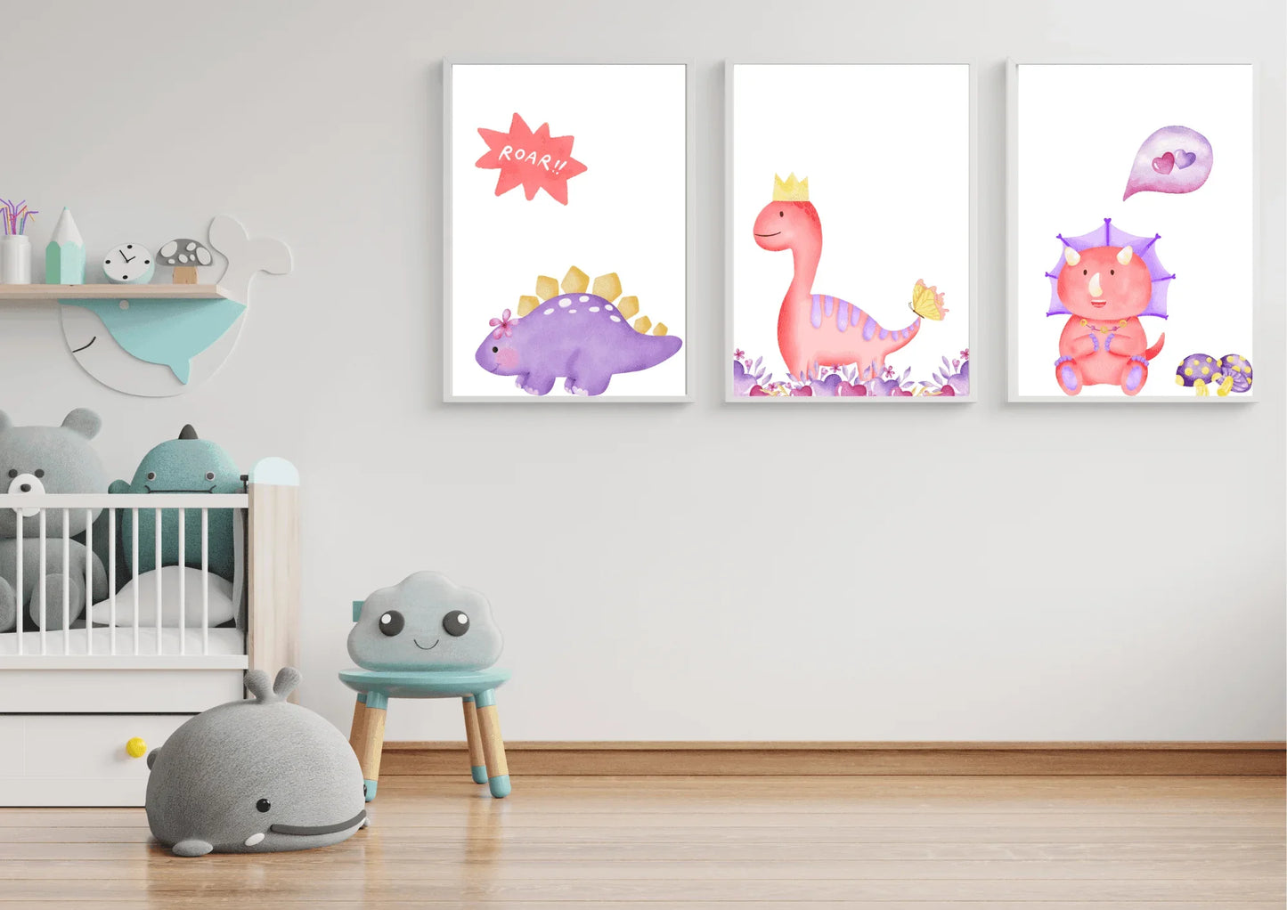 A trio of framed digital art prints featuring watercolor dinosaurs, hanging on a white wall in a minimalist nursery. The prints show a purple stegosaurus, a pink T-Rex with a crown, and a pink triceratops. Below the prints is a crib and a small blue chair with a smiling cloud face.