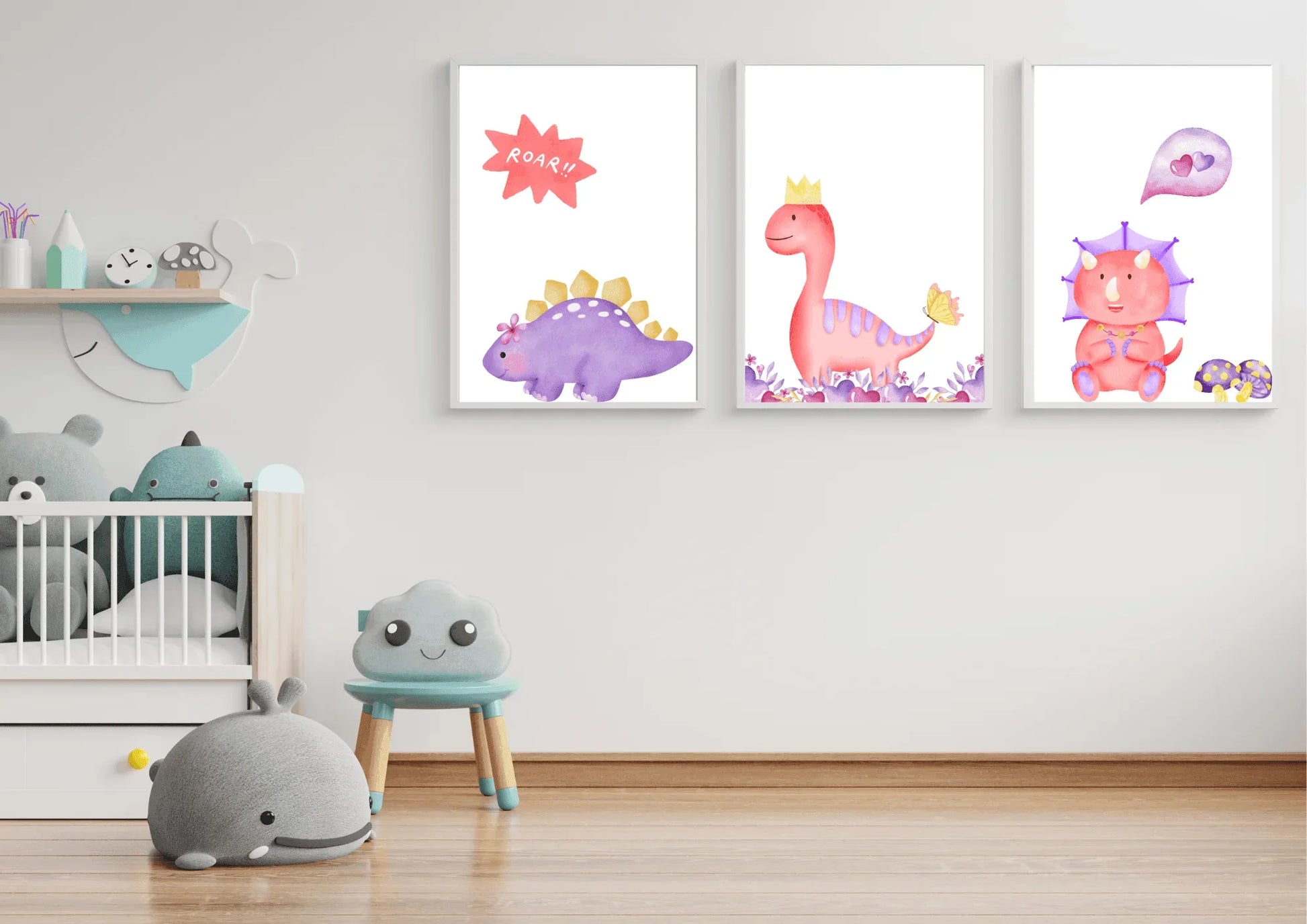 A trio of framed digital art prints featuring watercolor dinosaurs, hanging on a white wall in a minimalist nursery. The prints show a purple stegosaurus, a pink T-Rex with a crown, and a pink triceratops. Below the prints is a crib and a small blue chair with a smiling cloud face.