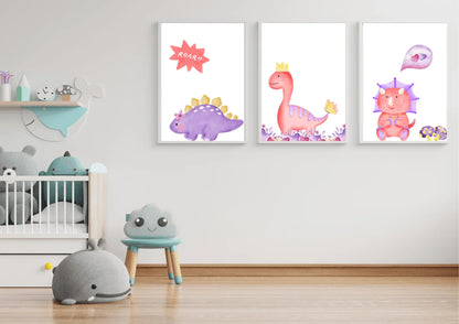 A trio of framed digital art prints featuring watercolor dinosaurs, hanging on a white wall in a minimalist nursery. The prints show a purple stegosaurus, a pink T-Rex with a crown, and a pink triceratops. Below the prints is a crib and a small blue chair with a smiling cloud face.