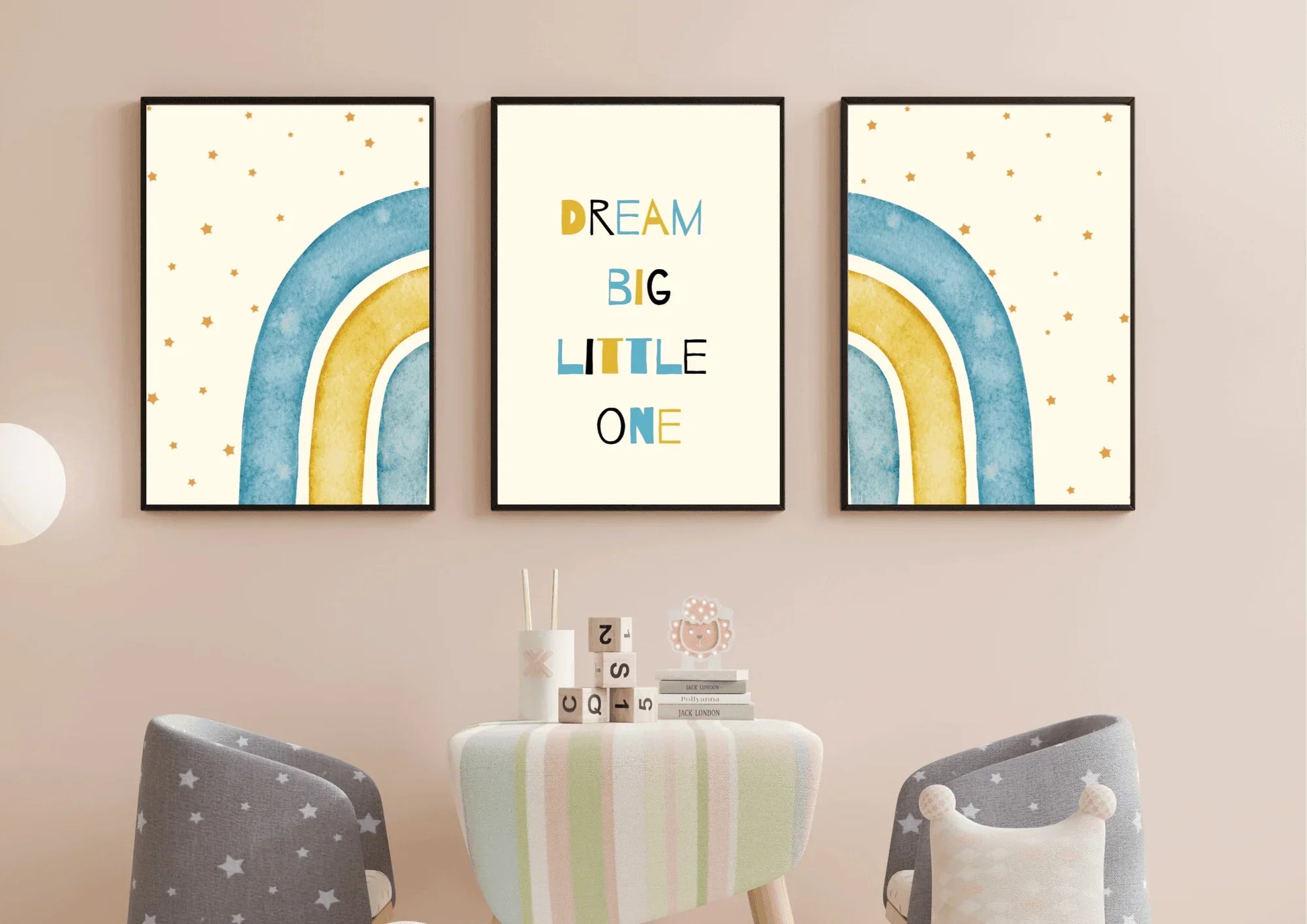 A three-piece framed nursery print set, featuring a "Dream Big Little One" message and blue and yellow watercolor rainbows, hanging on a light pink wall above a small table and two star-patterned chairs