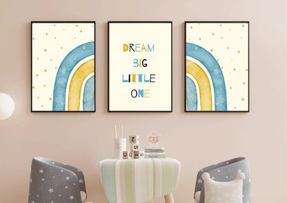 A three-piece framed nursery print set, featuring a "Dream Big Little One" message and blue and yellow watercolor rainbows, hanging on a light pink wall above a small table and two star-patterned chairs