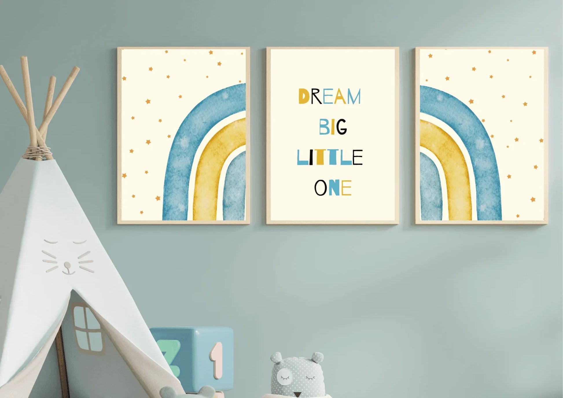 A three-piece framed nursery print set, featuring a "Dream Big Little One" message and blue and yellow watercolor rainbows, hanging on a light blue wall next to a white teepee.