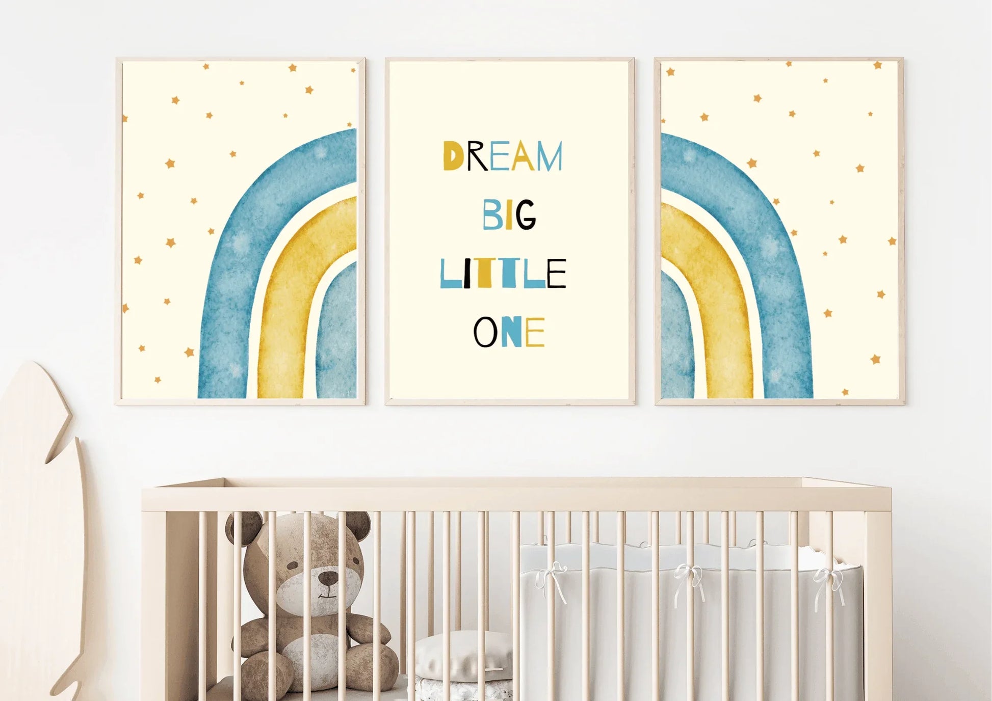 A three-piece framed nursery print set, featuring a "Dream Big Little One" message and blue and yellow watercolor rainbows, hanging on a white wall above a crib with a teddy bear.