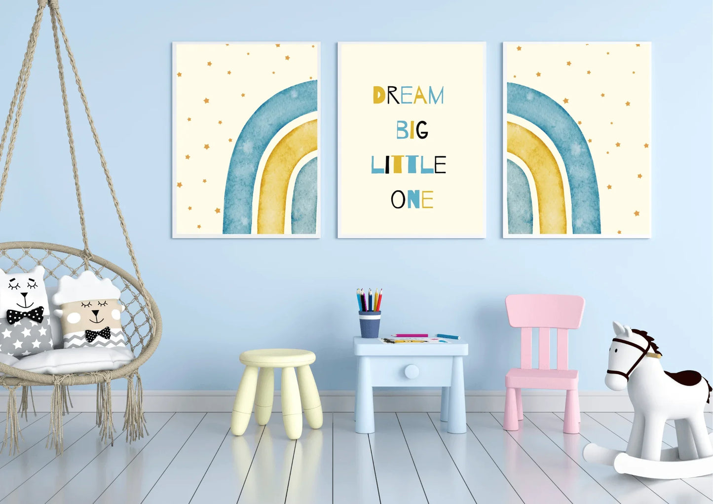 A three-piece framed nursery print set, featuring a "Dream Big Little One" message and blue and yellow watercolor rainbows, hanging on a light blue wall above a small table and a swing chair.