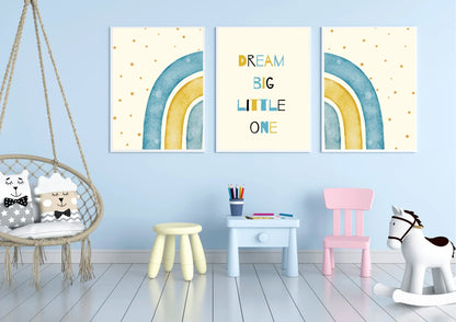 A three-piece framed nursery print set, featuring a "Dream Big Little One" message and blue and yellow watercolor rainbows, hanging on a light blue wall above a small table and a swing chair.