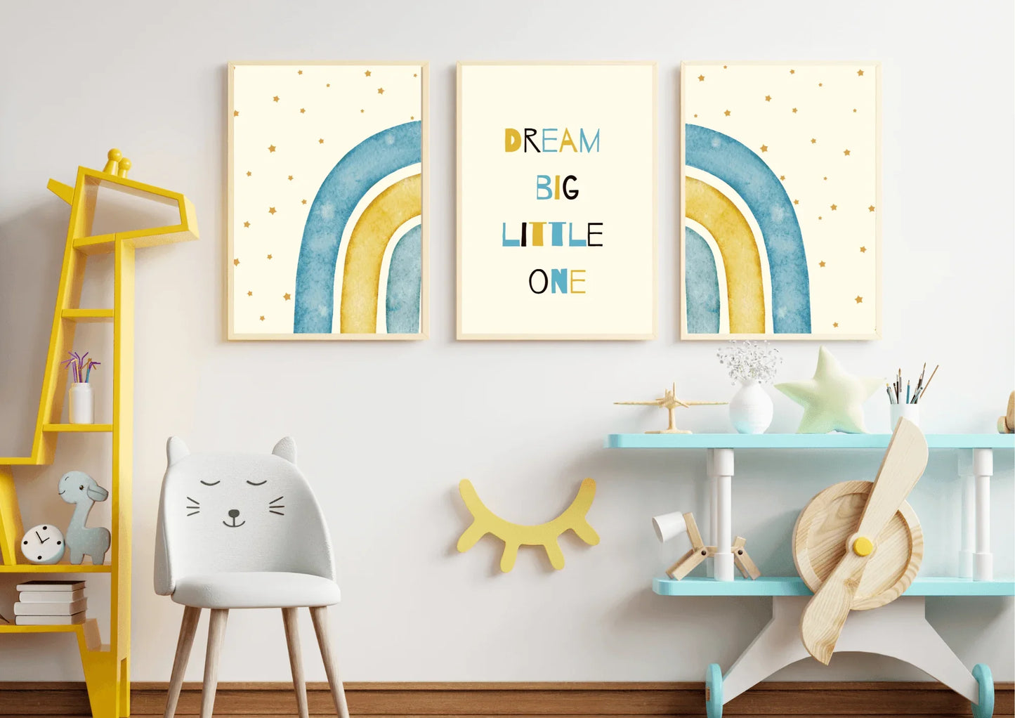 A three-piece framed nursery print set, featuring a "Dream Big Little One" message and blue and yellow watercolor rainbows, hanging on a white wall above a yellow bookshelf and a small chair.