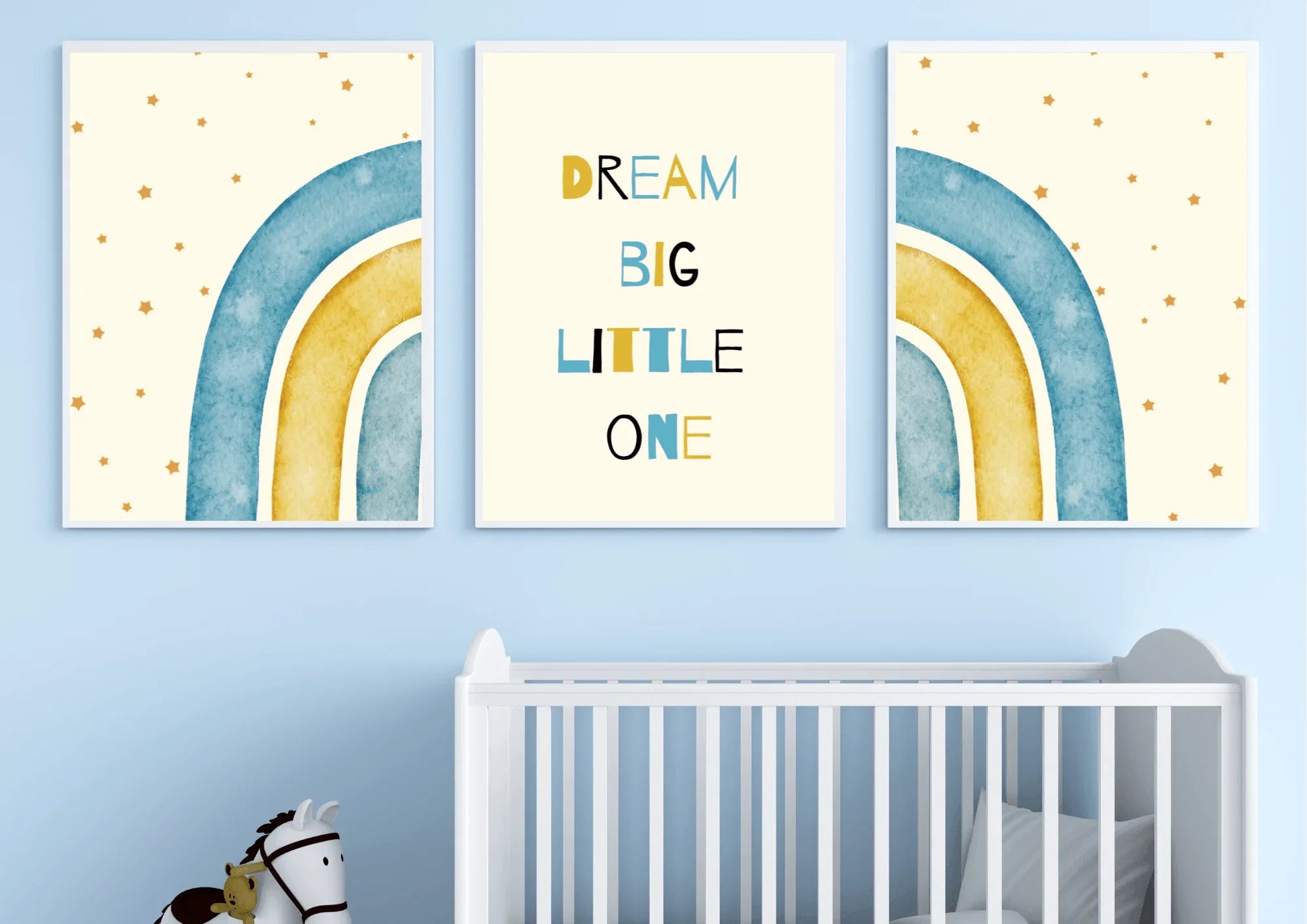 A three-piece framed nursery print set, featuring a "Dream Big Little One" message and blue and yellow watercolor rainbows, hanging on a light blue wall above a white crib.
