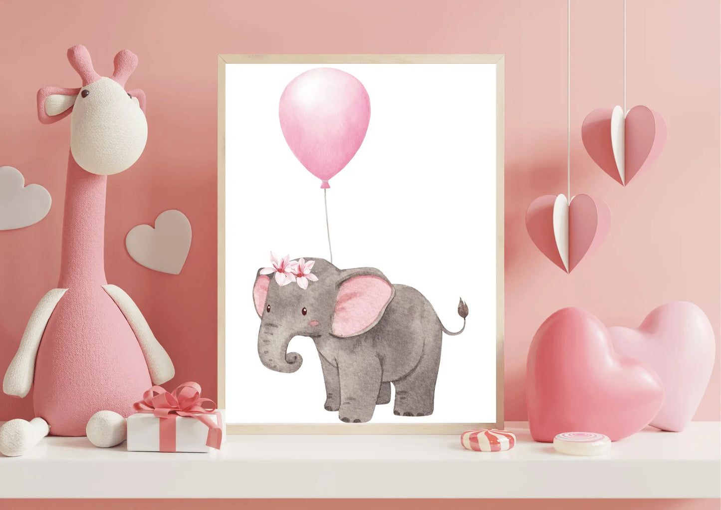 A framed nursery print of a baby elephant with a floral crown holding a pink balloon, standing on a white shelf next to a large stuffed giraffe and pink heart decorations.elephant nuresry print