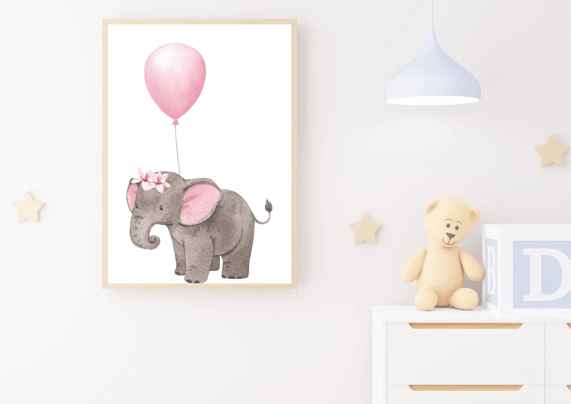A framed nursery print of a baby elephant with a floral crown holding a pink balloon, hanging on a white wall above a white dresser with a teddy bear and a lamp.