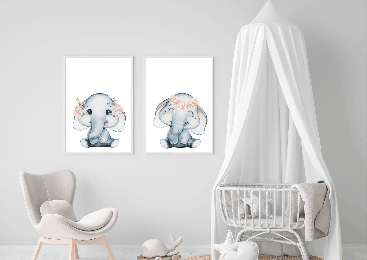 A bright, gender-neutral nursery room featuring a white wooden crib and natural wood furniture. Above the crib, two white-framed watercolor elephant prints are displayed side-by-side, creating a calming focal point. The elephants are soft grey with delicate pink flower crowns. The room includes a round woven jute rug, a rattan accent chair, and a small wooden side table with a floral arrangement. The light walls and abundance of natural wood create a peaceful, minimalist safari theme ideal for a baby's room