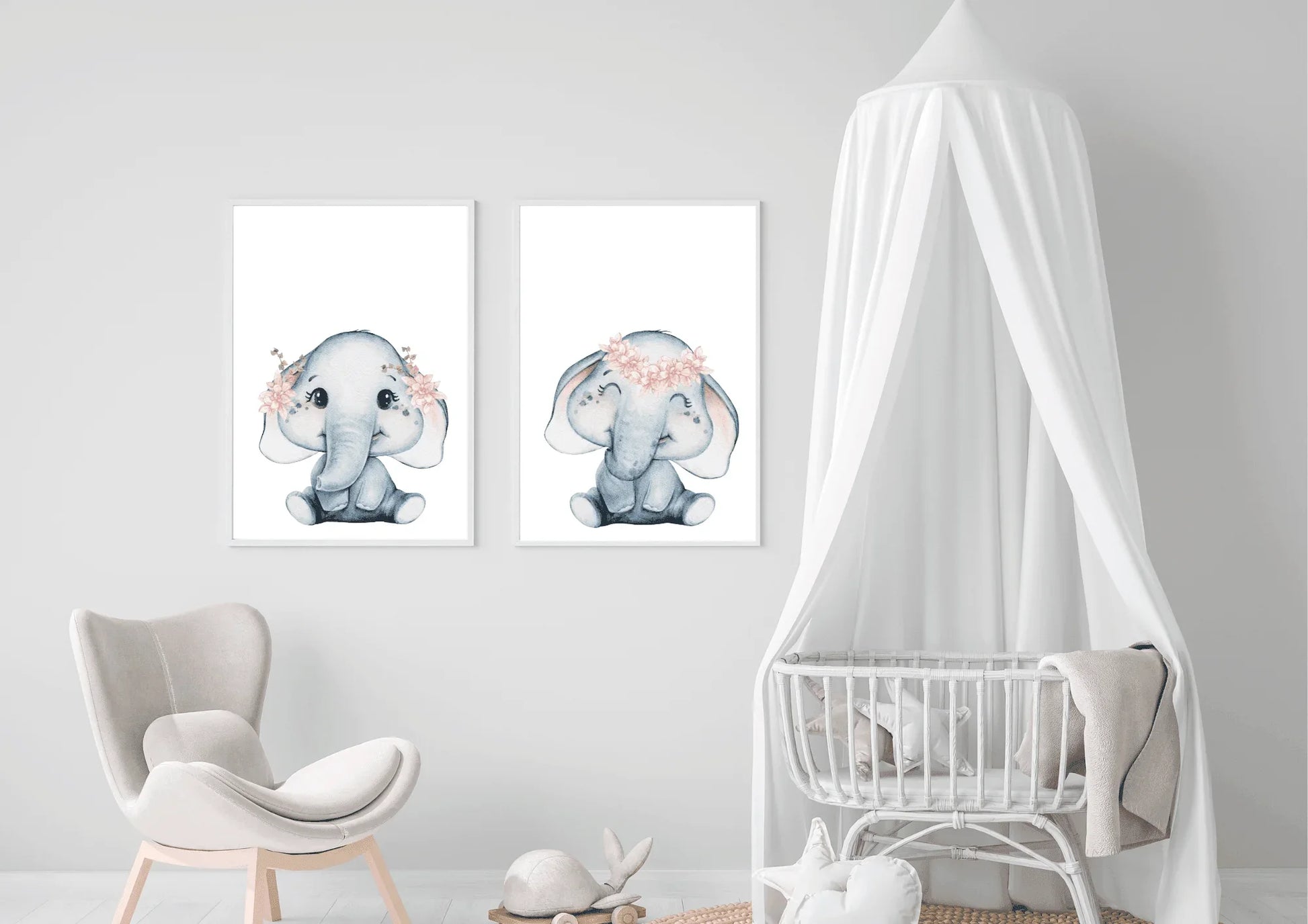 A bright, gender-neutral nursery room featuring a white wooden crib and natural wood furniture. Above the crib, two white-framed watercolor elephant prints are displayed side-by-side, creating a calming focal point. The elephants are soft grey with delicate pink flower crowns. The room includes a round woven jute rug, a rattan accent chair, and a small wooden side table with a floral arrangement. The light walls and abundance of natural wood create a peaceful, minimalist safari theme ideal for a baby's room