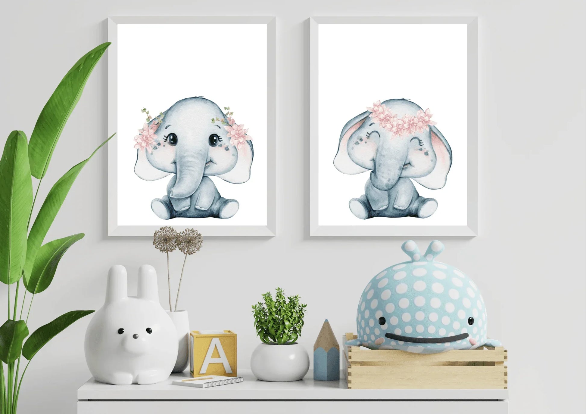 A close-up view of a natural wooden changing table topped with a white changing pad and a small, decorative potted plant. Hung directly above the change table are two white-framed watercolor elephant prints displayed side-by-side. The soft grey elephants are detailed with delicate pink flower crowns. The wall is painted a neutral light taupe or grey, creating a warm, earthy, and gender-neutral aesthetic focused on practical, high-quality nursery furniture.