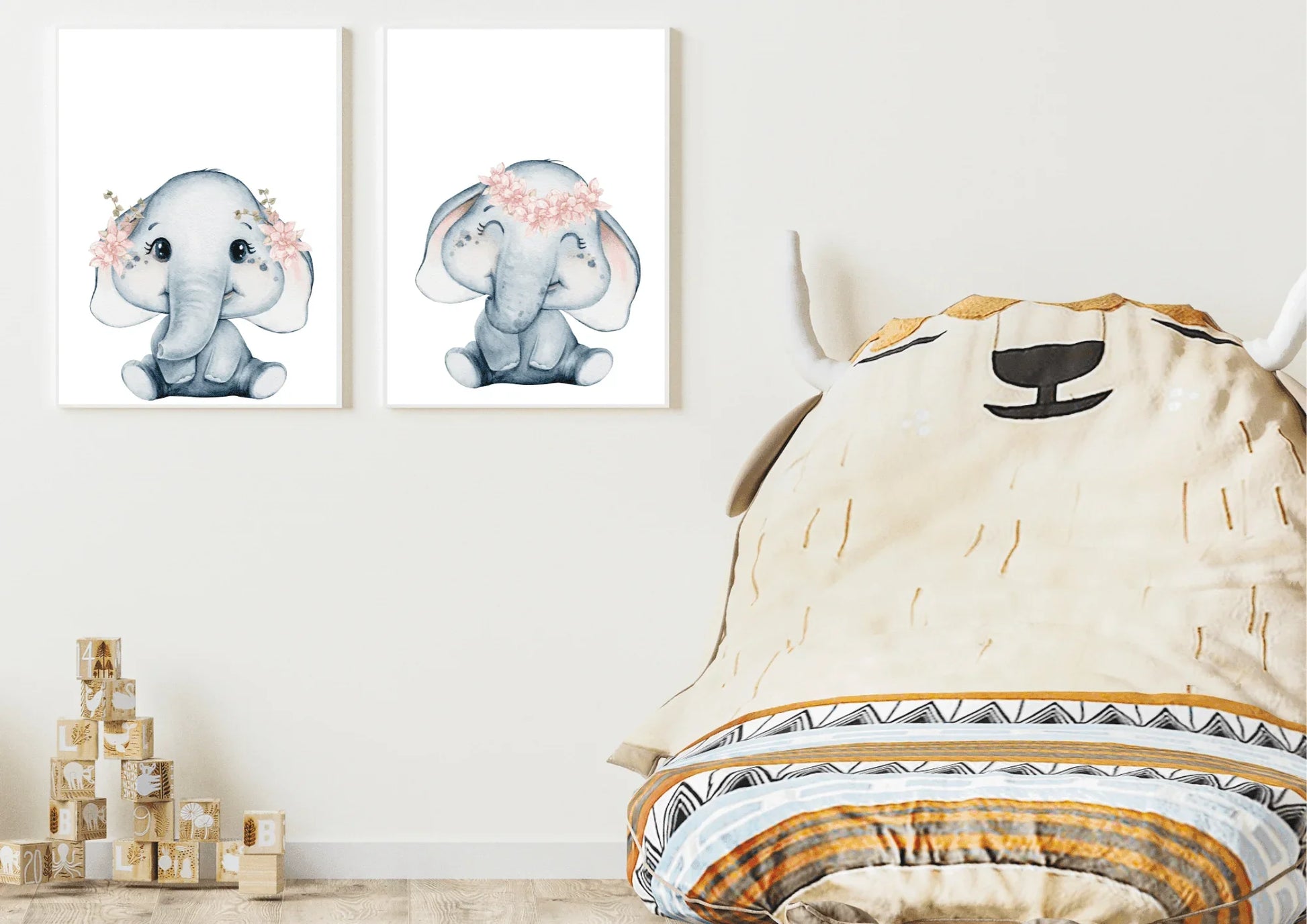 A close-up view focusing on a neutral, textured wall painted in a warm taupe or beige color. Hung directly above a row of natural wooden toy blocks are two white-framed watercolor elephant prints displayed side-by-side. The soft grey elephants are detailed with delicate pink flower crowns. The image emphasizes the gentle, gender-neutral color palette and the small, playful accents in the nursery decor.