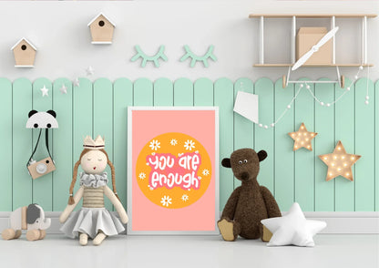 A framed "You are enough" art print on a two-toned wall in a child's playroom with a teddy bear and star lights.