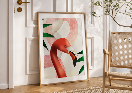 Framed wall art featuring a stylized pink flamingo illustration with green leaves, leaning against a white paneled wall in a sunlit room next to a woven chair.