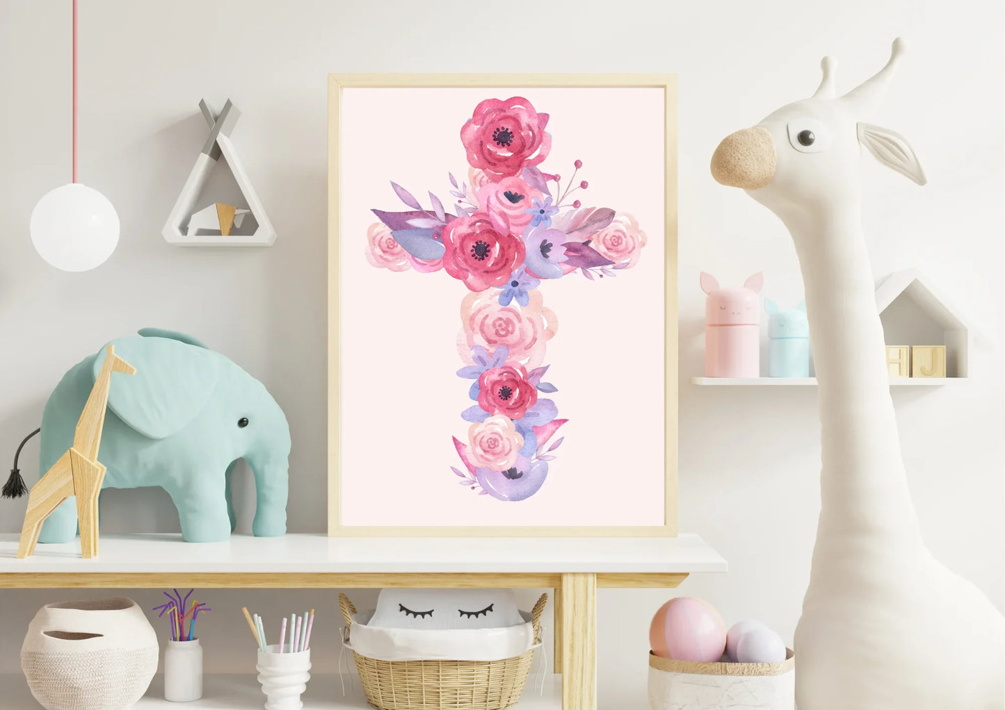 A framed art print of a cross made of pink and purple watercolor flowers, resting on a white shelf. On the shelf, there's a light blue toy elephant, a wooden teepee-shaped shelf with two small items, and a large toy giraffe head on the right.