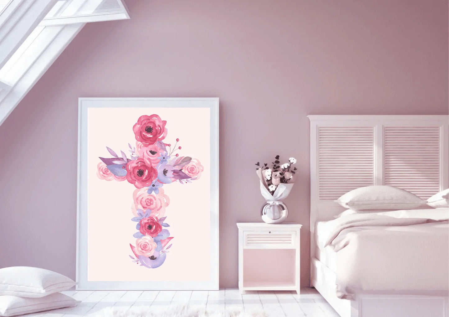 A bedroom with a slanted ceiling and pink walls. A large, framed art print of a cross made of pink and purple watercolor flowers leans against the wall. A white nightstand with a bouquet of flowers sits next to a bed with a white headboard and pink bedding.
