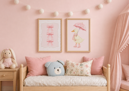 A cozy children's bedroom corner with a light wood daybed, pink pillows, a blue teddy bear pillow, a stuffed bunny on a side table, and two framed art prints on a pink wall. The prints show a pink bow and a duck with a pink umbrella.