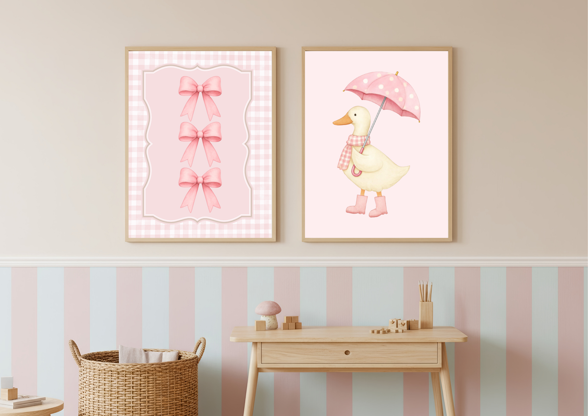 Two framed pink nursery wall art prints hanging above a light wood console table in a pastel room. The left print shows three pink bows on a pink gingham background, and the right print shows a yellow duck holding a pink umbrella with a pink scarf.