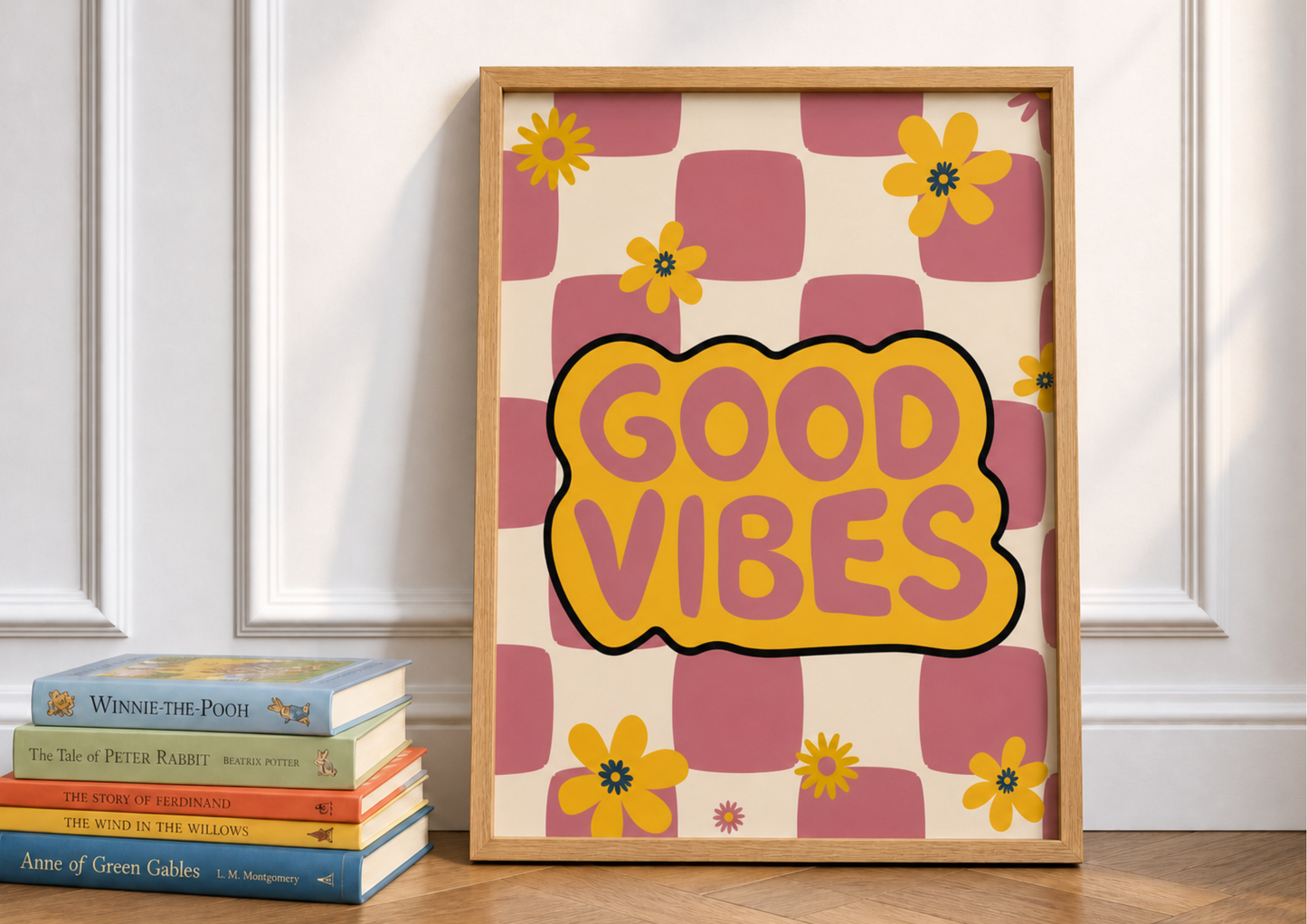 A framed retro-style art print leaning against a white paneled wall on a wooden floor, next to a stack of books. The print features a pink and cream checkered pattern with yellow flowers and a central yellow cloud shape outlined in black containing the words 'GOOD VIBES' in pink.