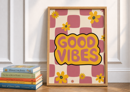 A framed retro-style art print leaning against a white paneled wall on a wooden floor, next to a stack of books. The print features a pink and cream checkered pattern with yellow flowers and a central yellow cloud shape outlined in black containing the words 'GOOD VIBES' in pink.