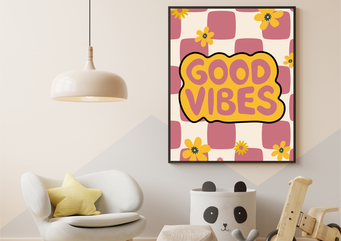 A framed wall art print with the words “GOOD VIBES” in yellow on a pink and cream checkered background with yellow flowers, shown hanging in a styled room with a white chair, pendant light, and wooden toy.