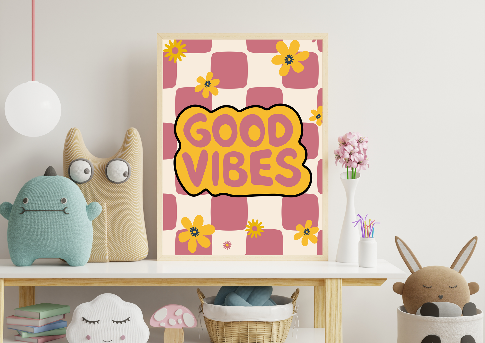 A framed art print with retro pink and yellow checkered squares and the words 'GOOD VIBES' in a yellow cloud shape, styled on a shelf in a cozy room setting with plush toys and flowers.