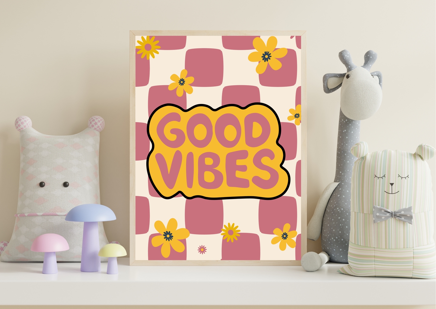 A framed art print with a retro pink and yellow checkerboard pattern, yellow flowers, and a central yellow cloud shape containing the text 'GOOD VIBES' in pink. The print is styled on a white shelf next to plush toys and small decorative mushrooms.