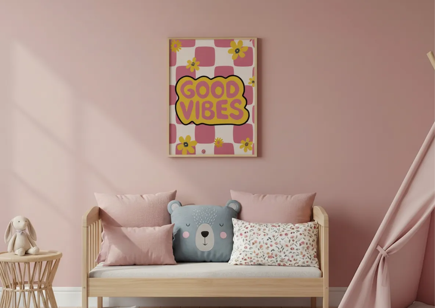 A framed wall art print with the words 'GOOD VIBES' in yellow on a pink patterned background, hanging above a light wood daybed with pink pillows in a soft pink nursery room.