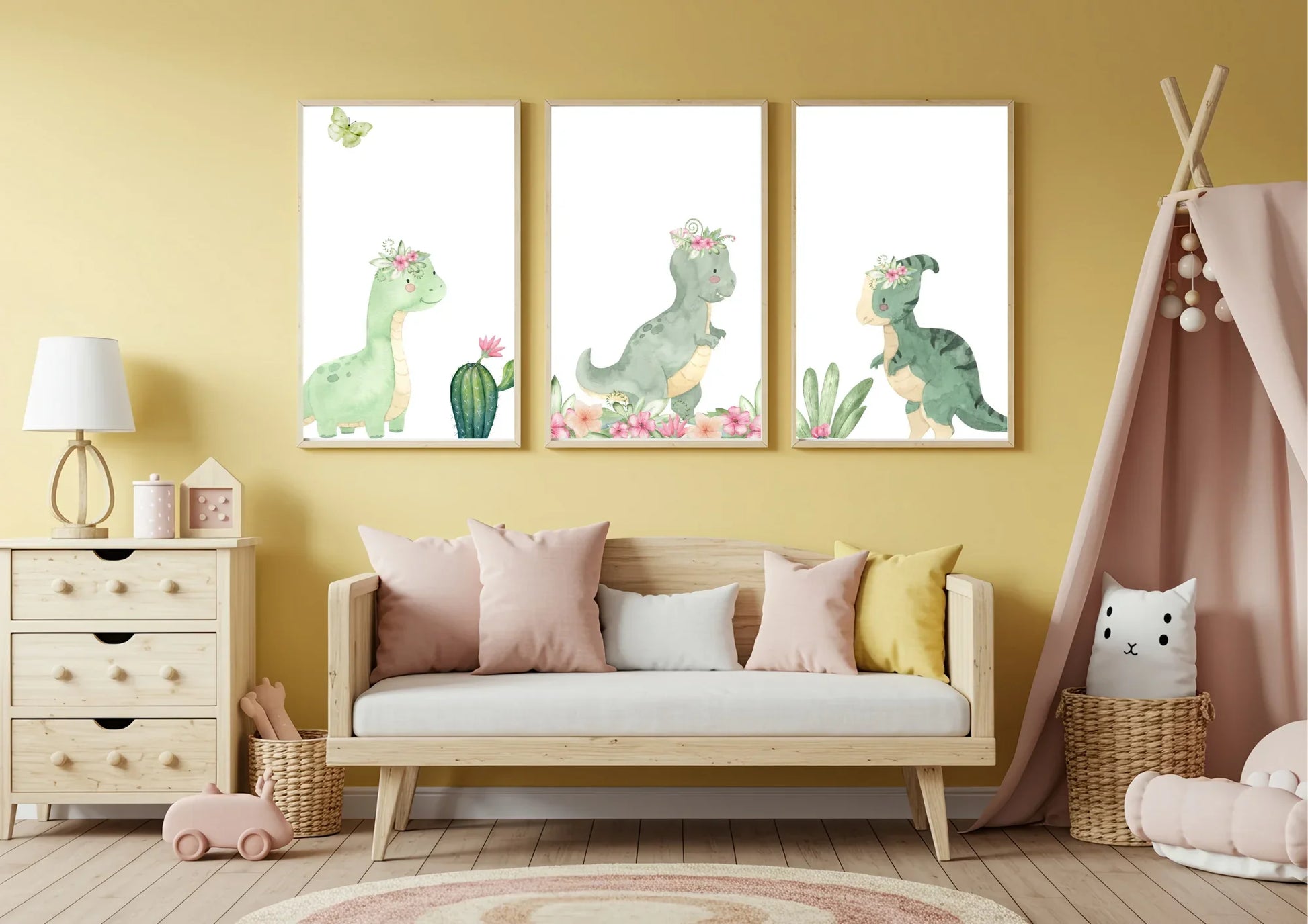 Three framed watercolor prints of cute green dinosaurs with floral crowns on a yellow nursery wall. The prints feature a long-necked dinosaur, a T-Rex, and a smaller dinosaur, each with plants or flowers. A daybed with pink, yellow, and white pillows sits below, with a wooden dresser to the left and a pink teepee to the right.